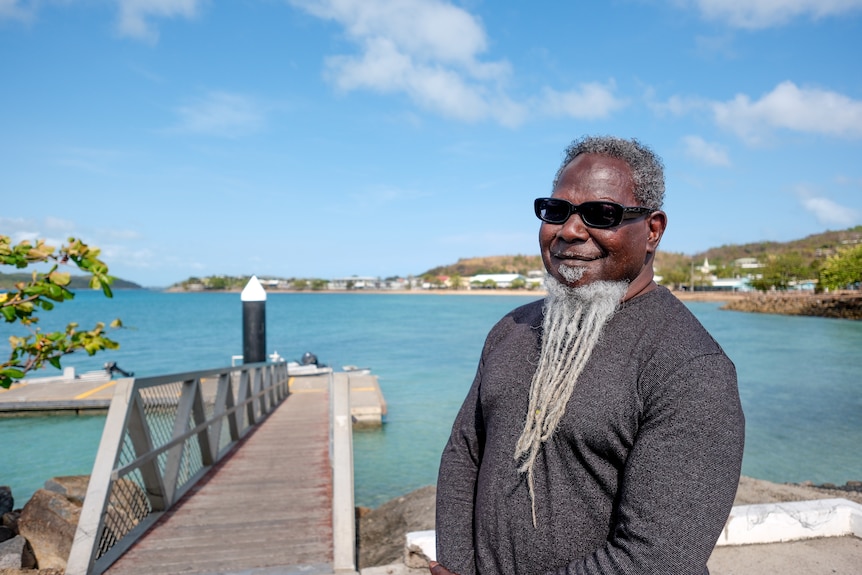 A man wearing a dark jumper and sunglasses with a long grey beard stands beside a wharf. 