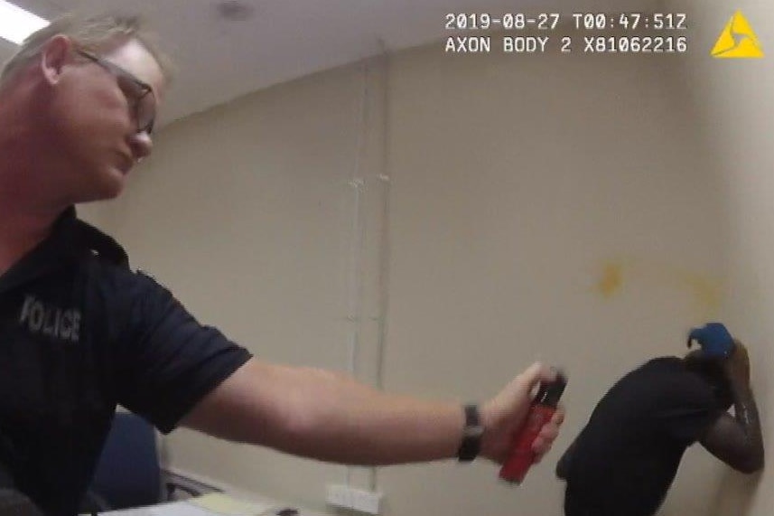 A police officer can be seen using pepper spray Aboriginal man in a prison cell. The man is covering his face.