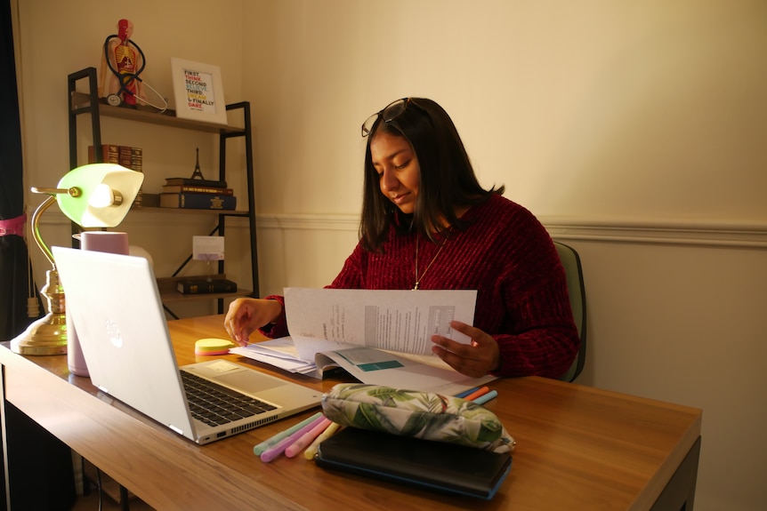 A young Indian woman studying in her room. There's a lamp and laptop and papers on her table. She is looking down and smiling.
