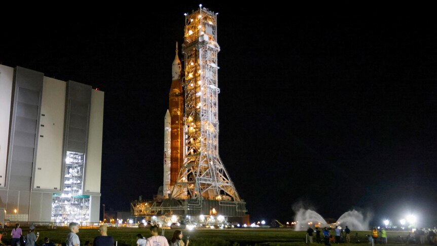 People sit on a lawn at night as a space rocket surrounded by scaffolding stands tall