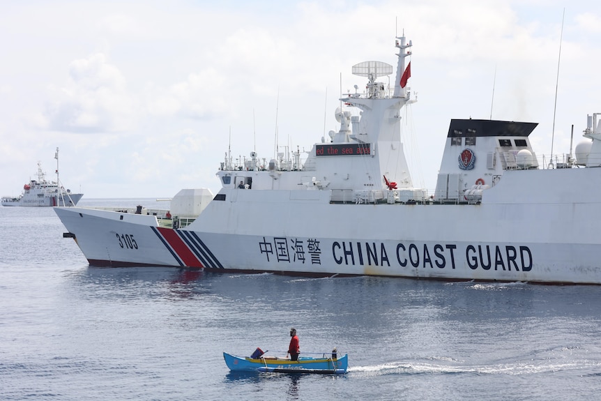 a fisherman, stands in his tiny wooden boat near a large China Coast Guard ship