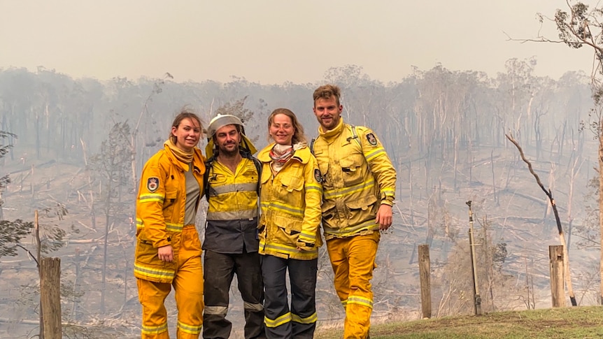 firefighters stand arm in arm on a green patch of hill surrounded by still smoking burnt forest