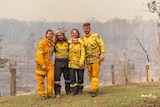 firefighters stand arm in arm on a green patch of hill surrounded by still smoking burnt forest