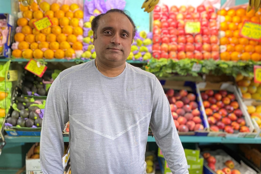 An Indian man in a grey long sleeve t-shirt stands at the counter in his fruit shop looking into the camera.