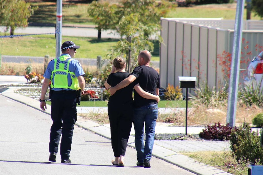 A couple walk with their arms around each other along a street accompanied by a police officer.