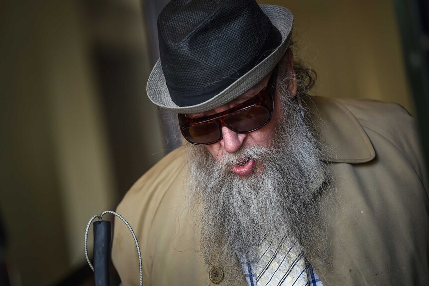 Robin Fletcher with a large beard, wearing a hat, sunglasses and carrying a cane.