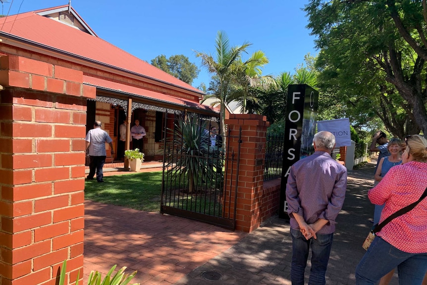 People stand outside the gates of a house for auction at Clarence Park. A large 'FOR SALE' sign is erected nearby.