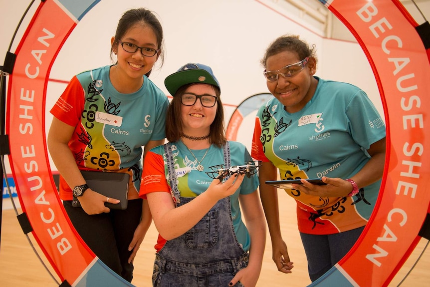A group of three teenage girls pose with a drone inside one of the obstacle course loops they have learned to fly through.