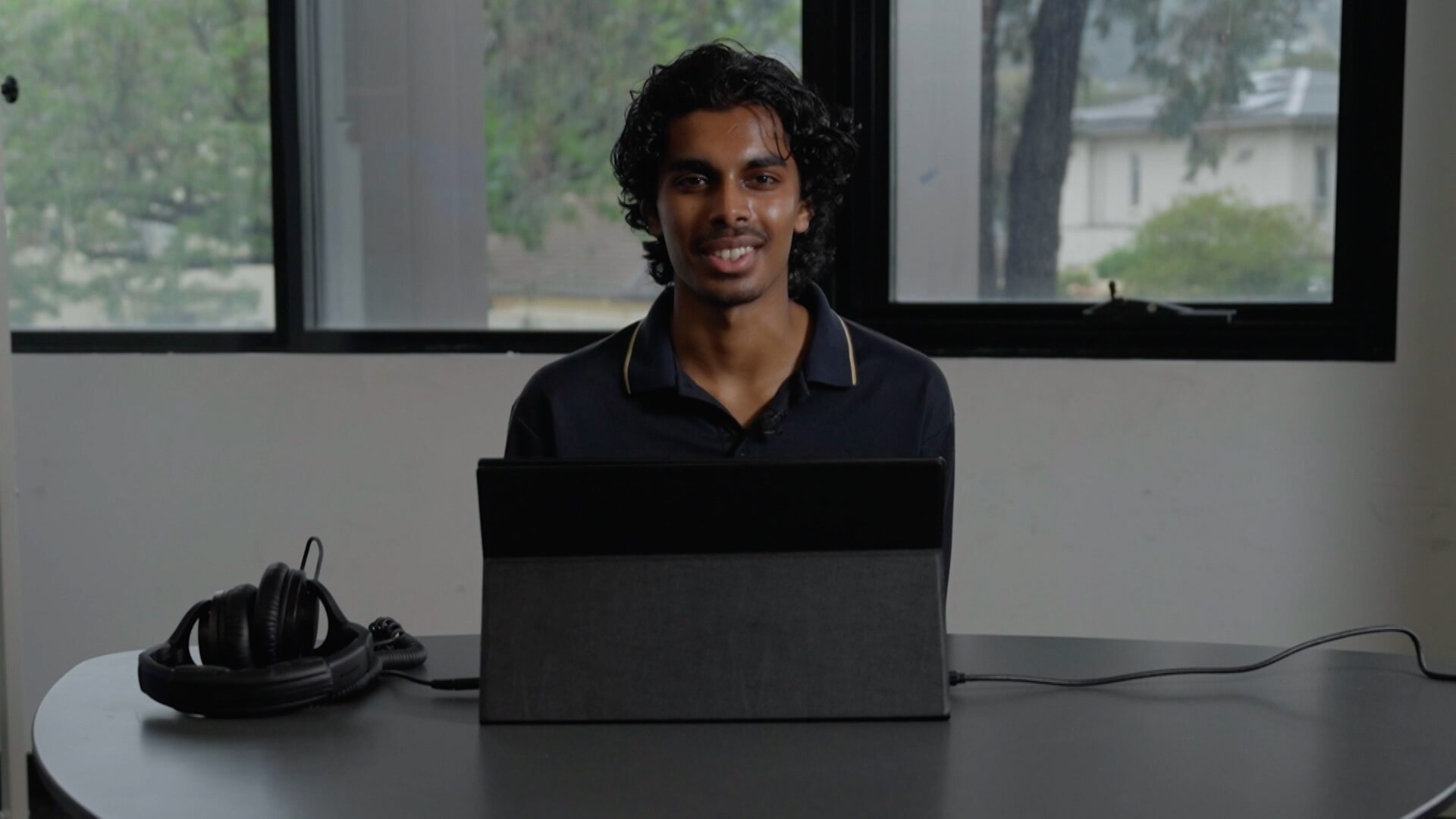 A young man with dark hair, wearing a dark shirt, sits smiling in front of a desk and computer. A window is in the background.
