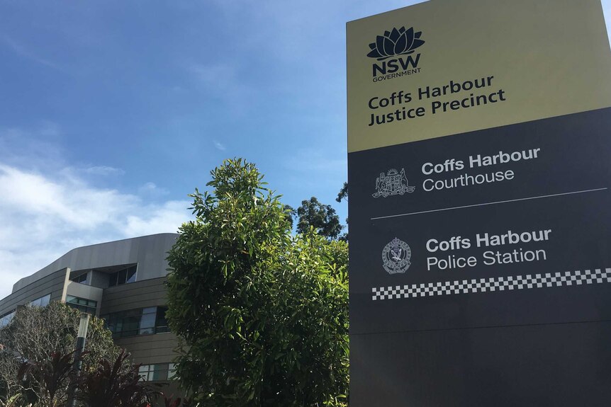 A tall sign in front of a tree and a large building. The sign reads: Coffs Harbour Justice Precinct.