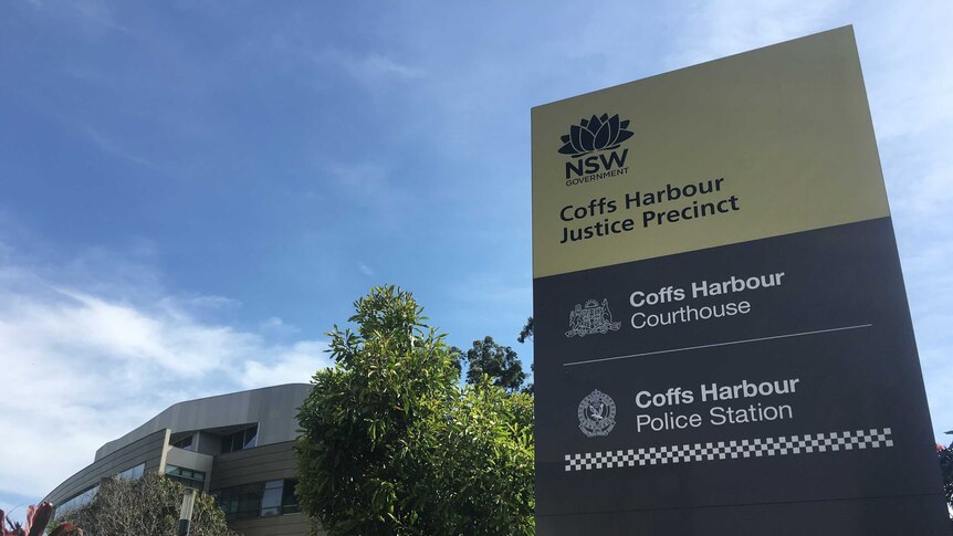 A tall sign in front of a tree and a large building. The sign reads: Coffs Harbour Justice Precinct.