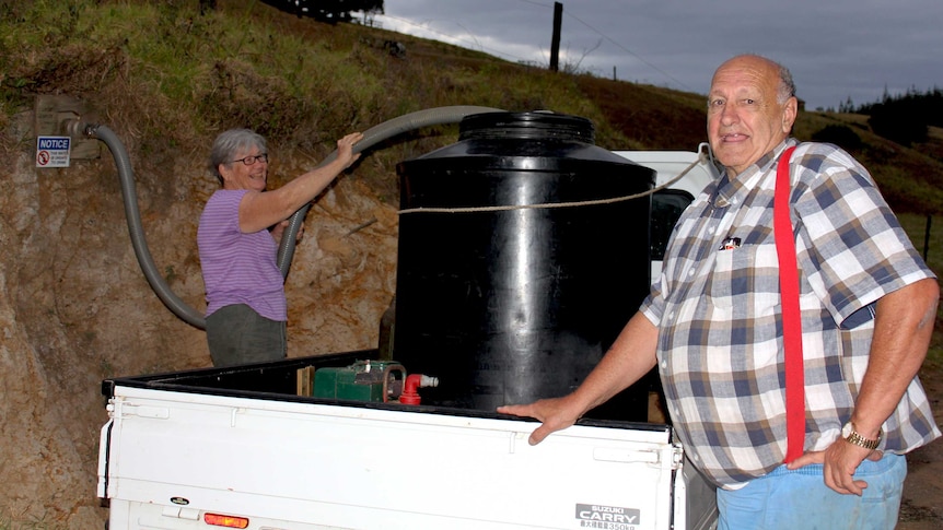 A man and a woman stand next to a ute while as they fill a water tank with a hose connected to a natural spring