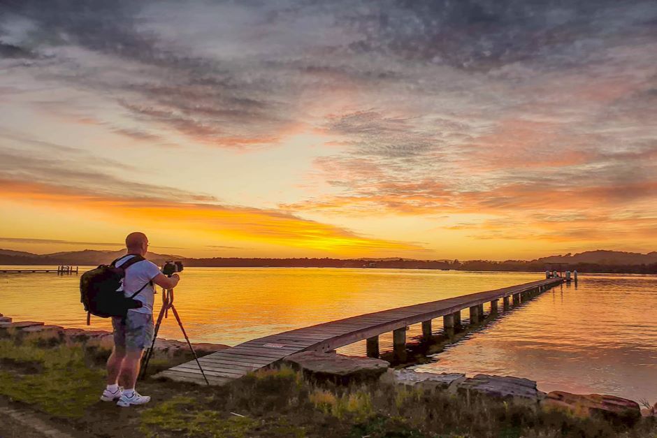 Man with camera and tripod standing at end of jetty at sunset.