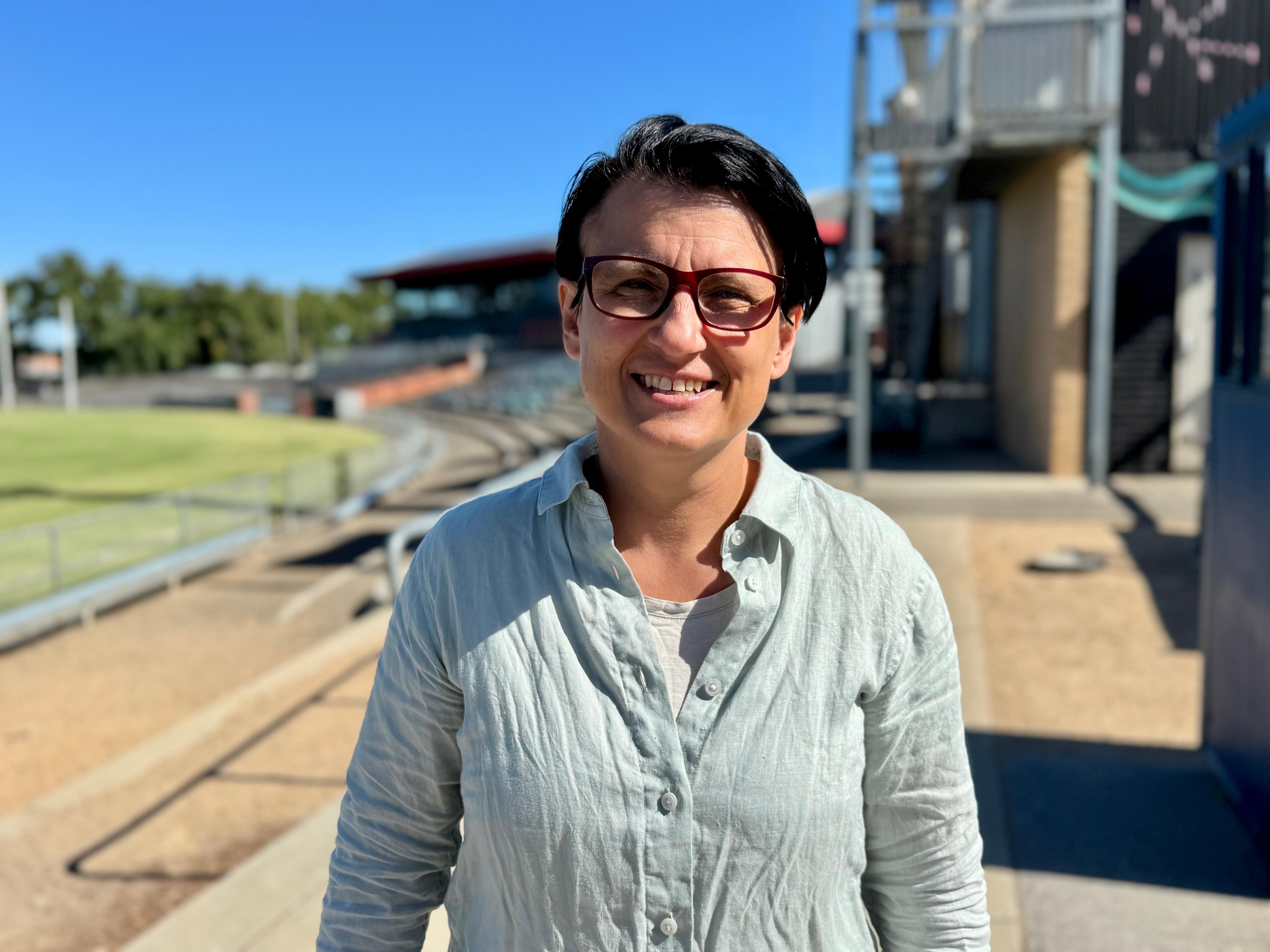 Una mujer con rasgos faciales oscuros y cabello corto sonríe a la cámara cerca de Footy Oval 