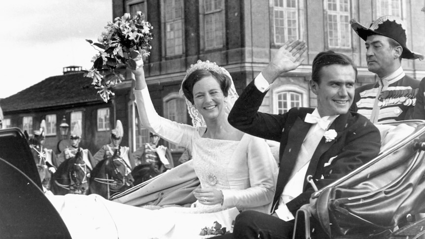 A bride and groom smile from the back of an open carriage, waving and holding a bouquet aloft