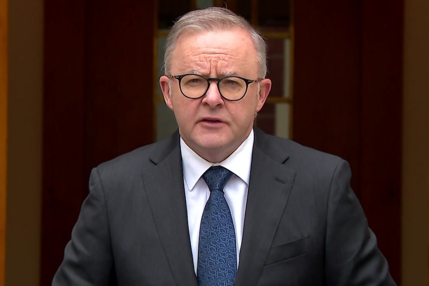 A man in glasses and a suit stands between Aboriginal and Torres Strait Islander flags.