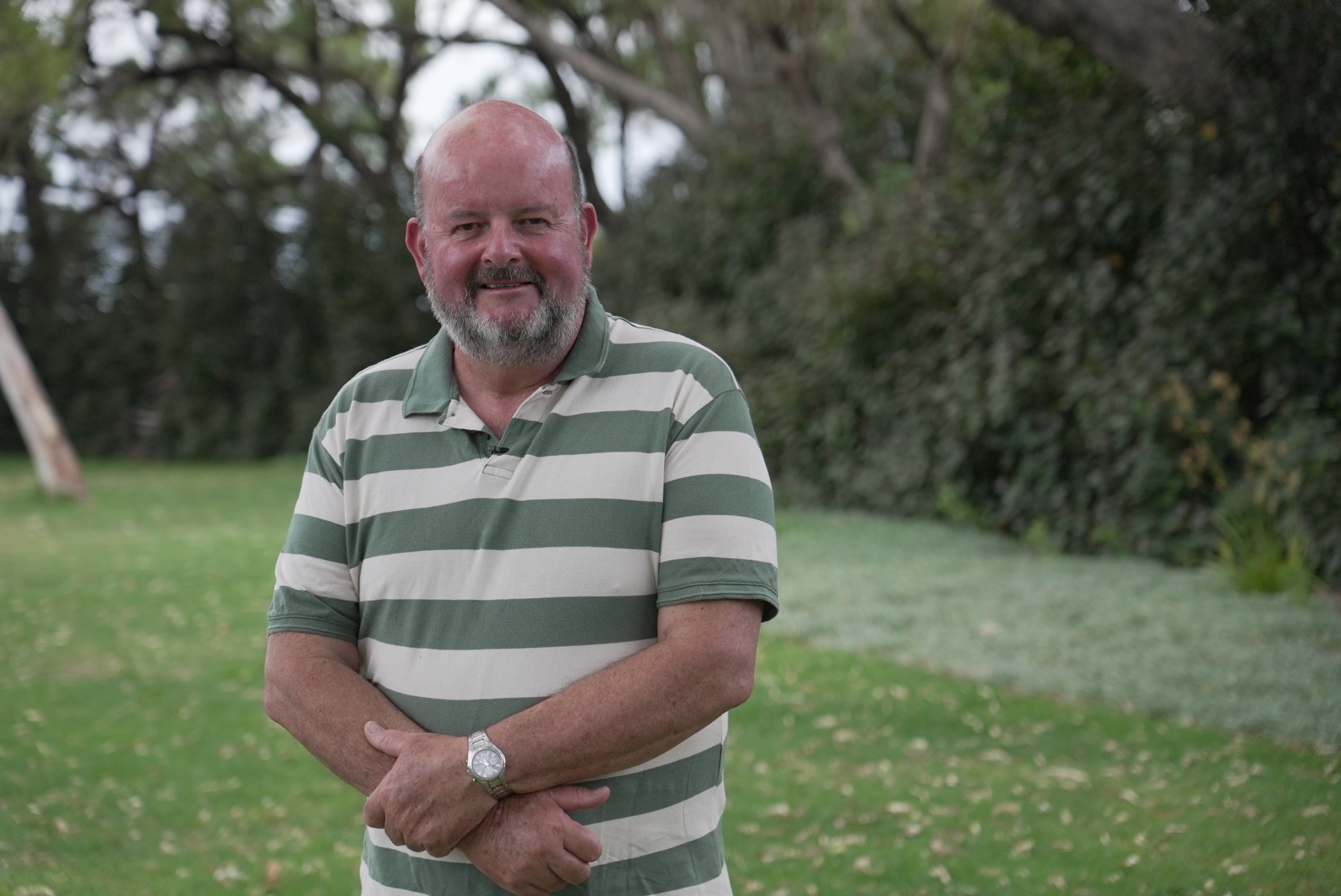 Balding man with a beard wearing a white and green striped shirt, standing in a park with arms folded