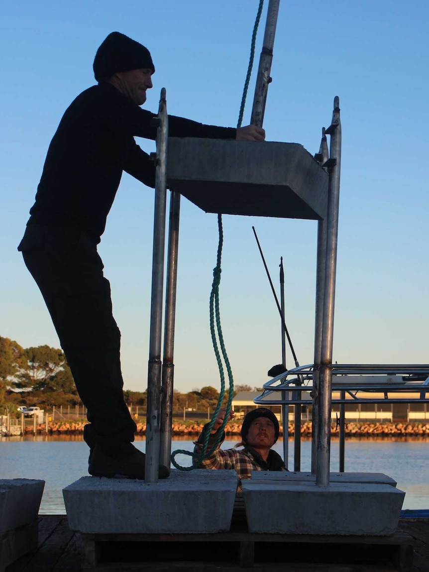 A man is silhouetted on a trailer, guiding a concrete block as a crane lifts it