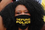 A woman raises her hand in protest while wearing a 'defund the police' mask
