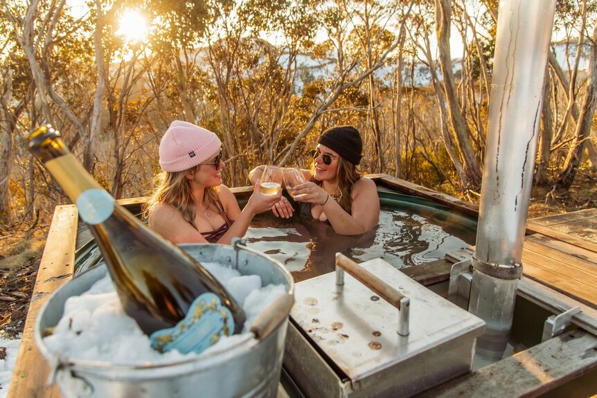 Two women clink wine glasses in a hot tub outside. 