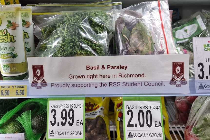 Basil and parsley on a supermarket shelf with a sign indicating the produce was grown at the local state school