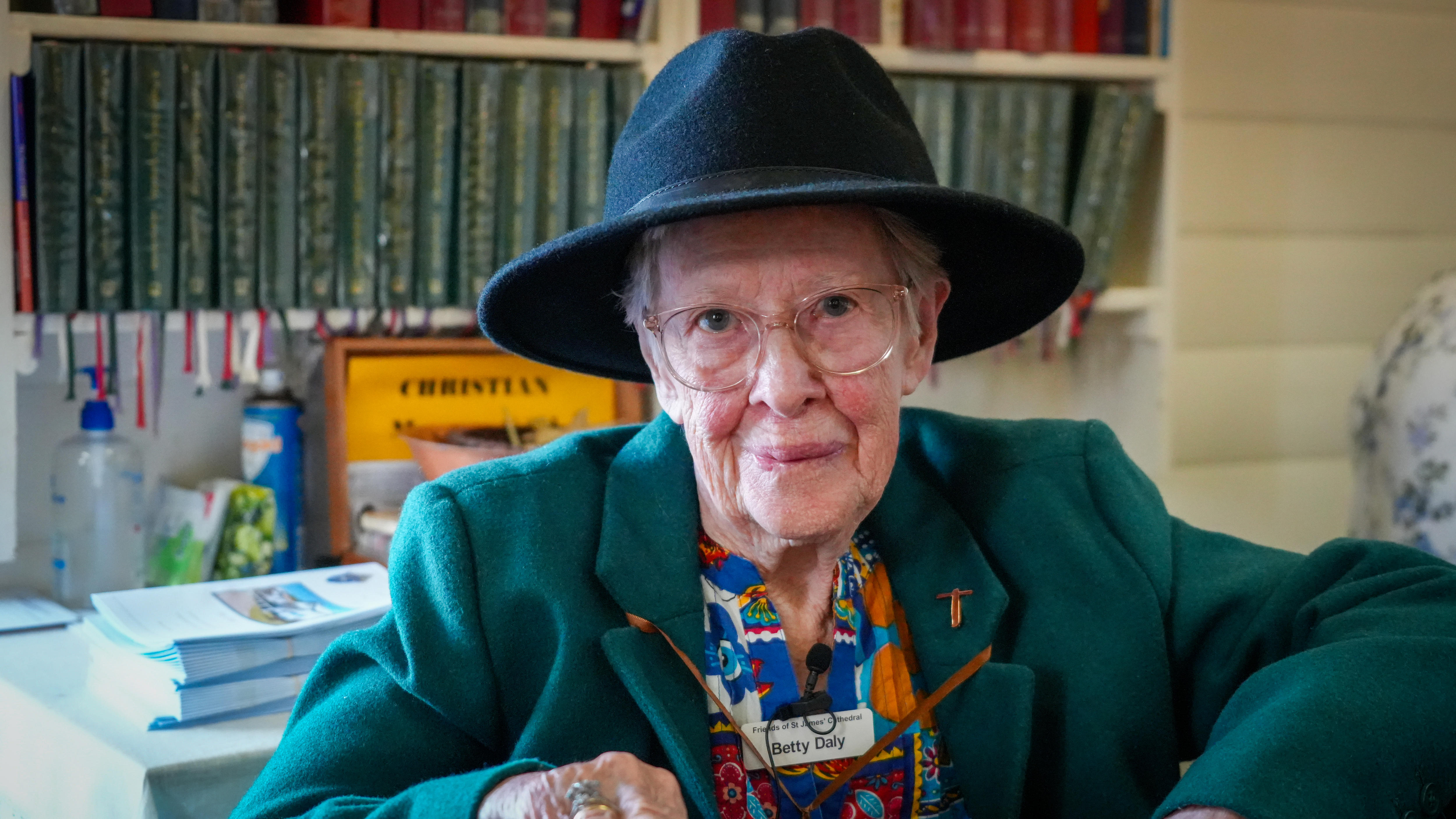 An elderly woman sits in a chair, wearing a black hat and a green velvet jacket