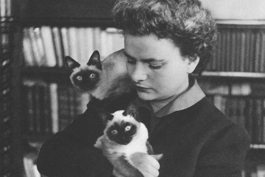 A black and white photo of Elsa Morante, posing in front of a bookshelf with two Siamese cats