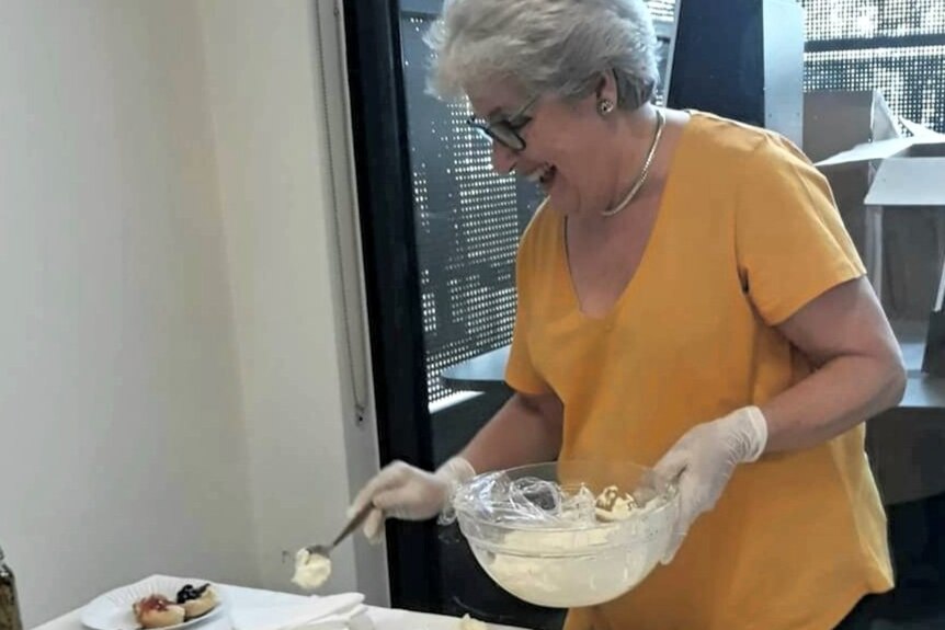 An older woman smiles broadly as she dollops cream onto some scones.
