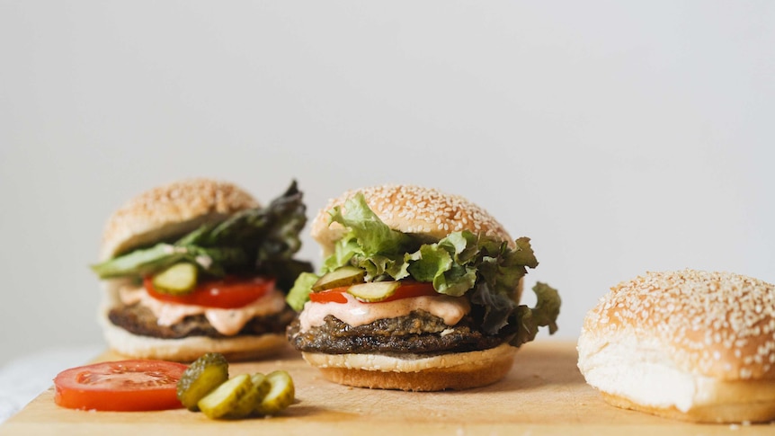 Two vegetable burgers on a chopping board with cheese-filled mushrooms, pickles, tomato slices, fresh lettuce and soft buns.