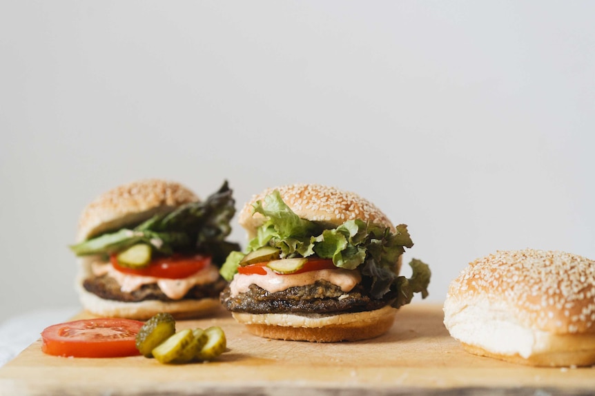 Two vegetable burgers on a chopping board with cheese-filled mushrooms, pickles, tomato slices, fresh lettuce and soft buns.