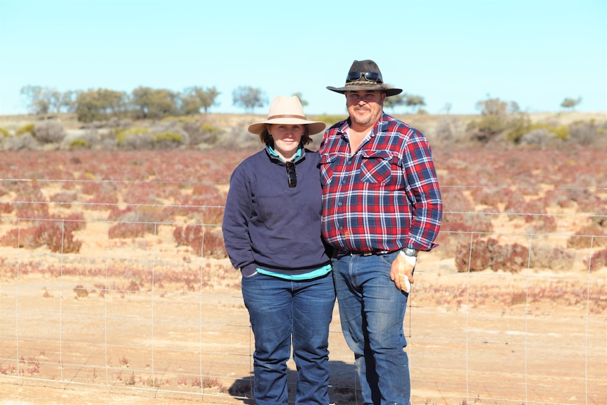 A woman and man stand side by side with an arid landscape behind them. They are wearing broad brim hats and long sleeve shirts.