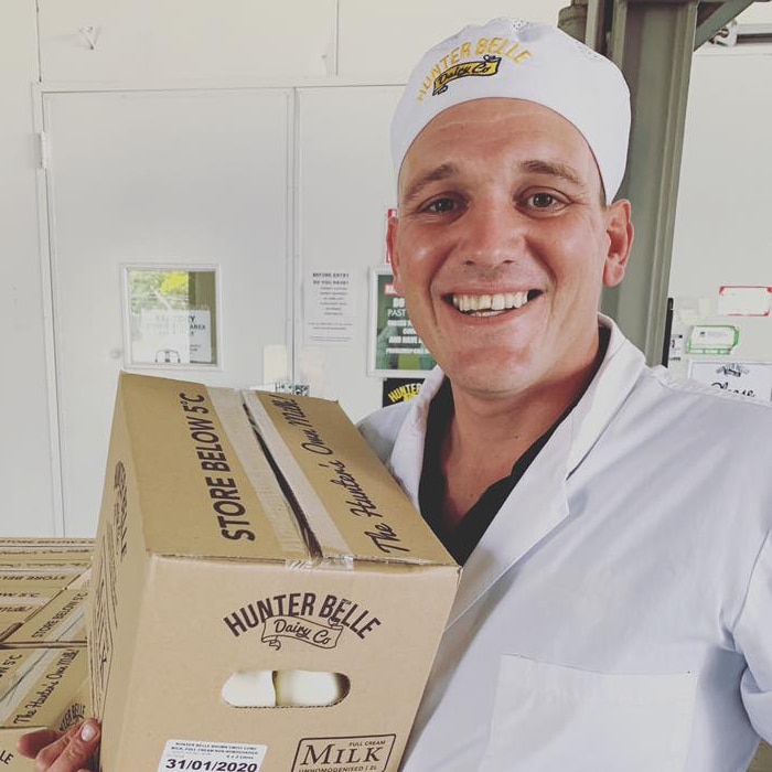 A man in a white coat and hat holds a box of 2 litre milk bottles. He is smiling.