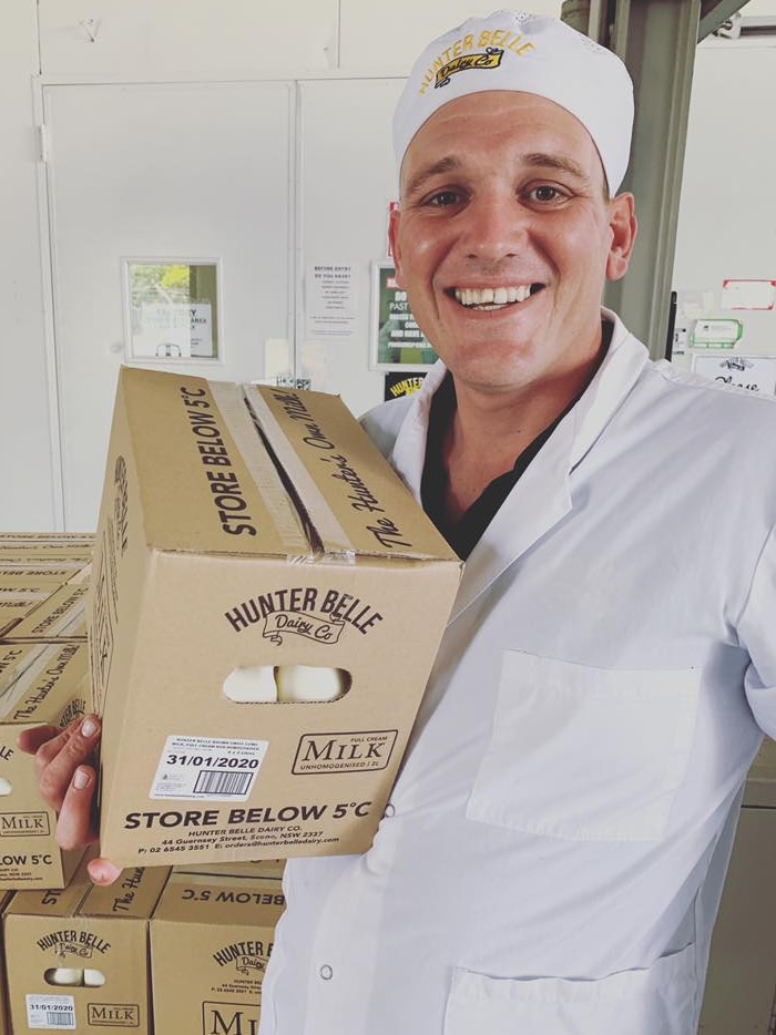 A man in a white coat and hat holds a box of 2 litre milk bottles. He is smiling.