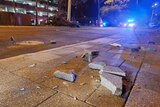Broken paving stones on a footpath with a police car in the background with its lights on at night.
