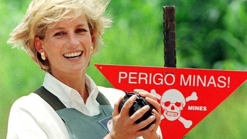 Blonde white woman wears white shirt and protective vest smiles and holds what looks like a land mine in front of warning sign