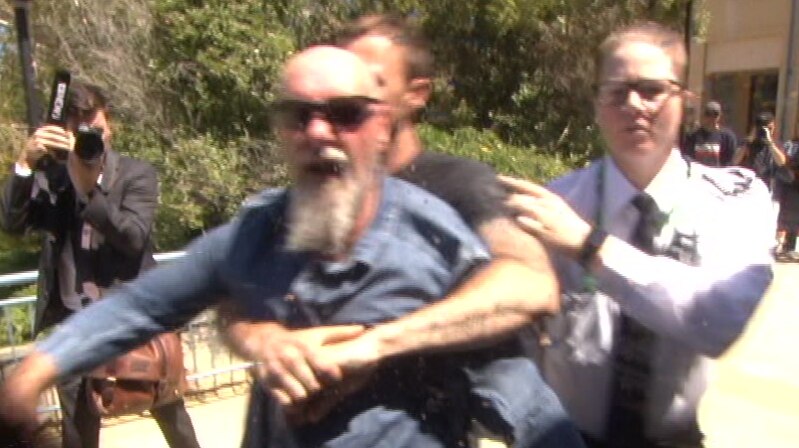 A man in a blue shirt and sunglasses aims a punch at a camera operator while being restrained by another man.