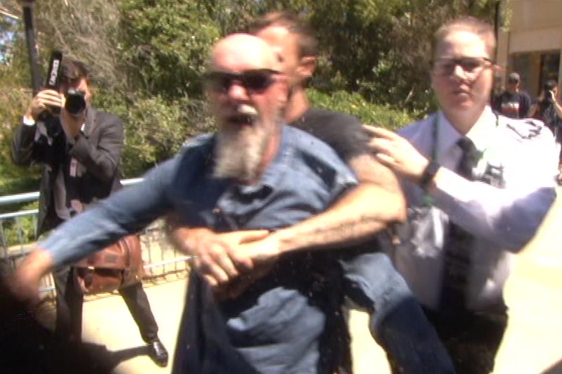 A man in a blue shirt and sunglasses aims a punch at a camera operator while being restrained by another man.