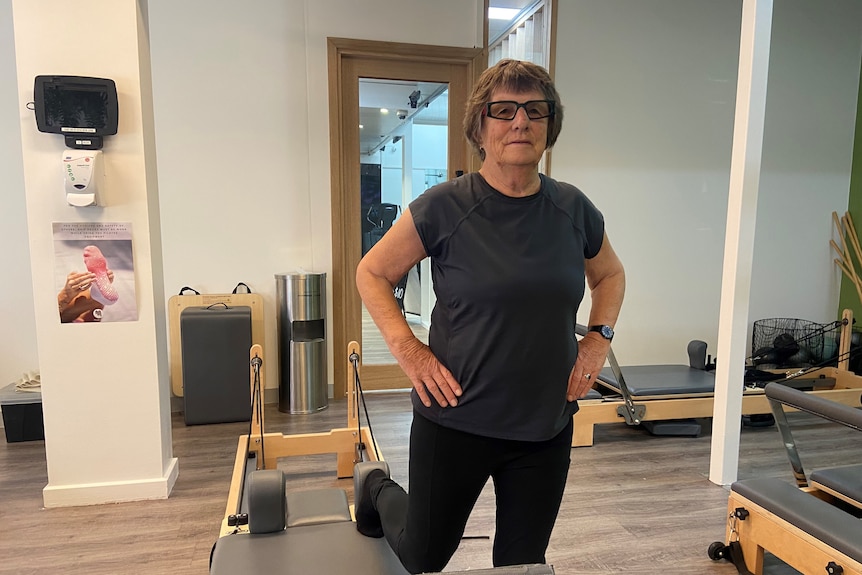 An older, bespectacled woman stretching in a Pilates studio.