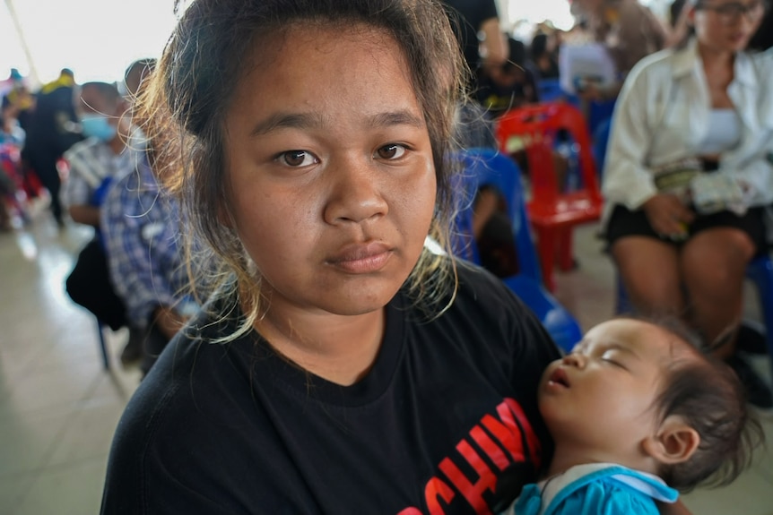 A young woman cradling a sleeping baby looks directly into camera