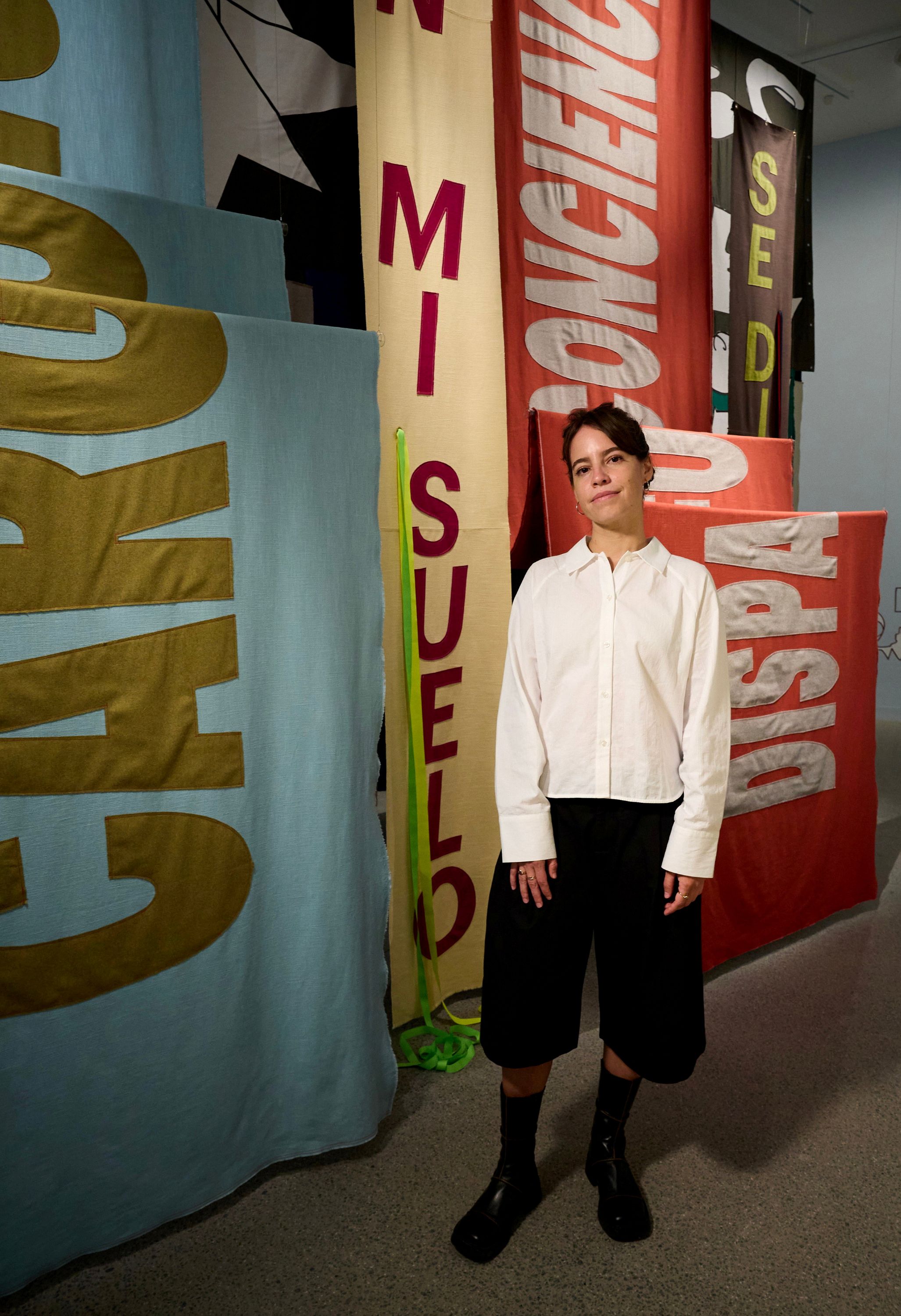 A young woman with short dark hair, wearing a white shirt and black pants, standing in front of textile banners.