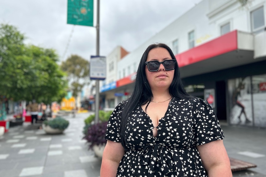 Woman in a black dress with printed white florals wearing sunglasses, standing on a street.