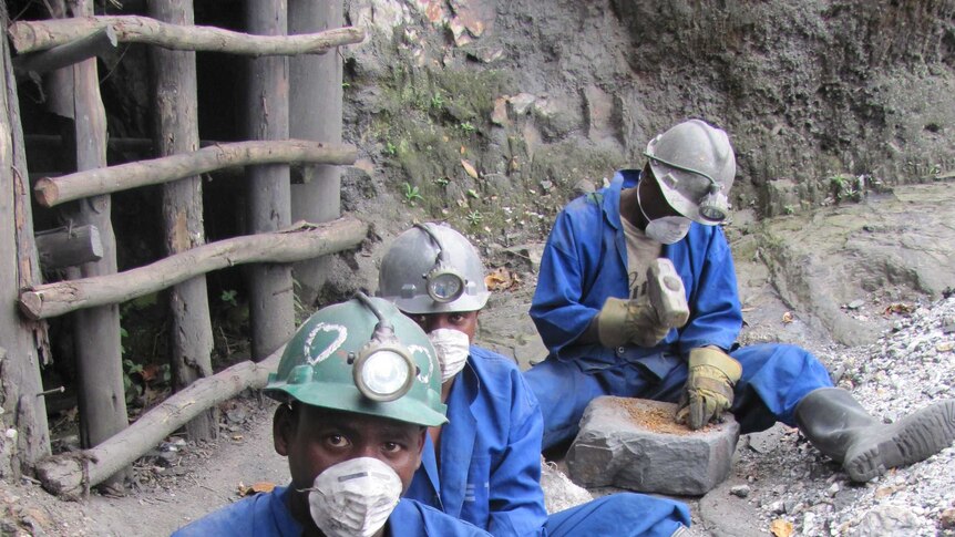 Three African miners sitting down and using mallets to pound large rocks to get minerals to make coltan