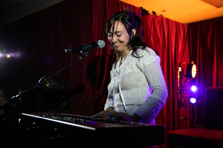 An Indigenous woman in her 20s smiles brightly as she performs behind a keyboard