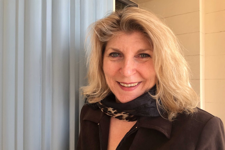 A woman with shoulder-length blonde hair, wearing a black scarf, smiles in front of a wall and pillar.