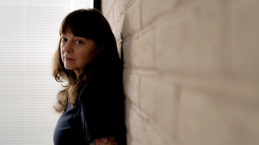 A woman with a tattoo sleeve leans against a white brick wall and looks to camera