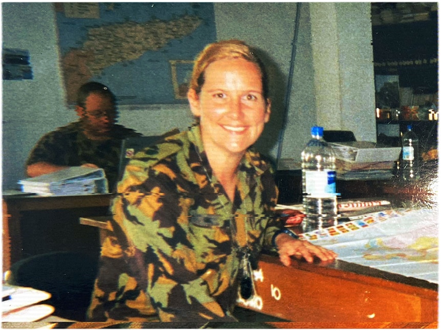 A woman in an army uniform sits at a desk turning to smile at the camera. A map sits before her, another map is on the wall.