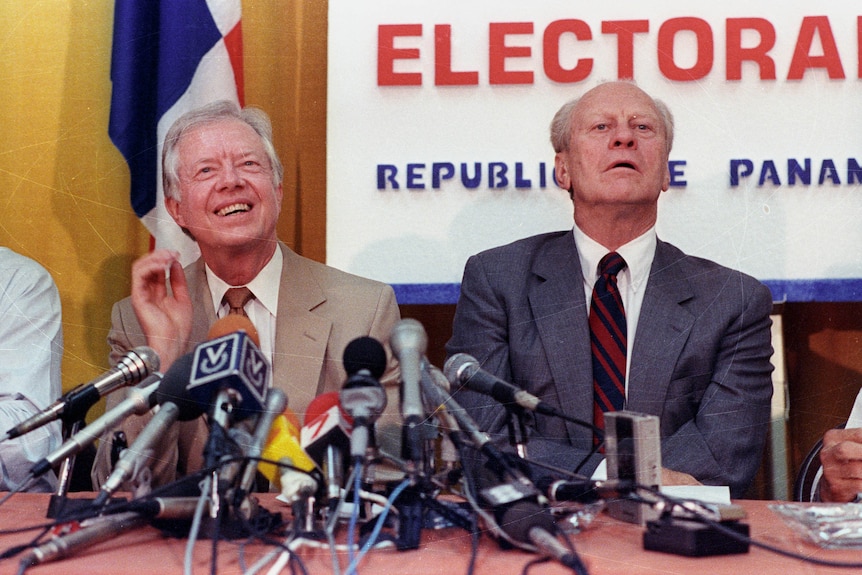Two men sit at a table with microphones in front of them. 