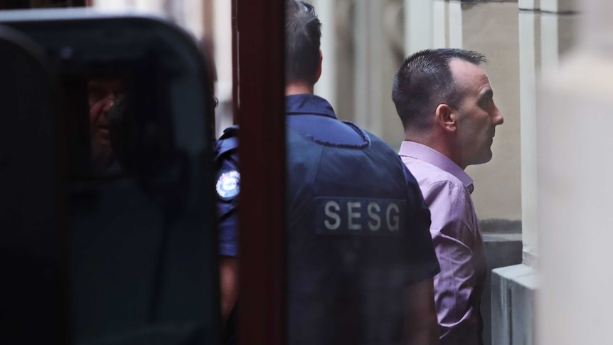 Jason Roberts arrives at the Supreme Court of Victoria wearing a pink collared shirt, accompanied by an SESG officer.