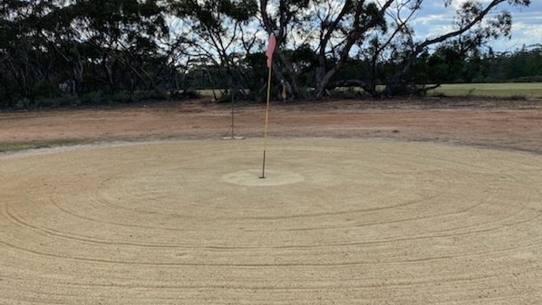 A raked sand scrape around a golf hole with mallee scrub and a green fairway in background.