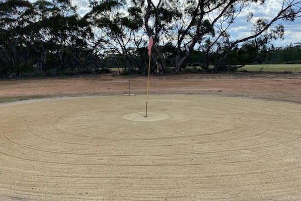 A raked sand scrape around a golf hole with mallee scrub and a green fairway in background.